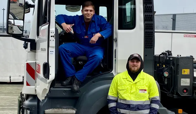 Ein Berufskraftfahrer sitzt mit der Tür geöffnet im Fahrzeug und ein anderer steht in gelber Sicherheitskleidung davor.
